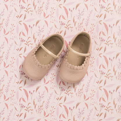Soft pink baby shoes with scalloped edges and a Mary Jane strap on patterned pink background.