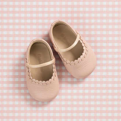 Soft pink baby shoes with scalloped edges and Mary Jane strap on gingham background.