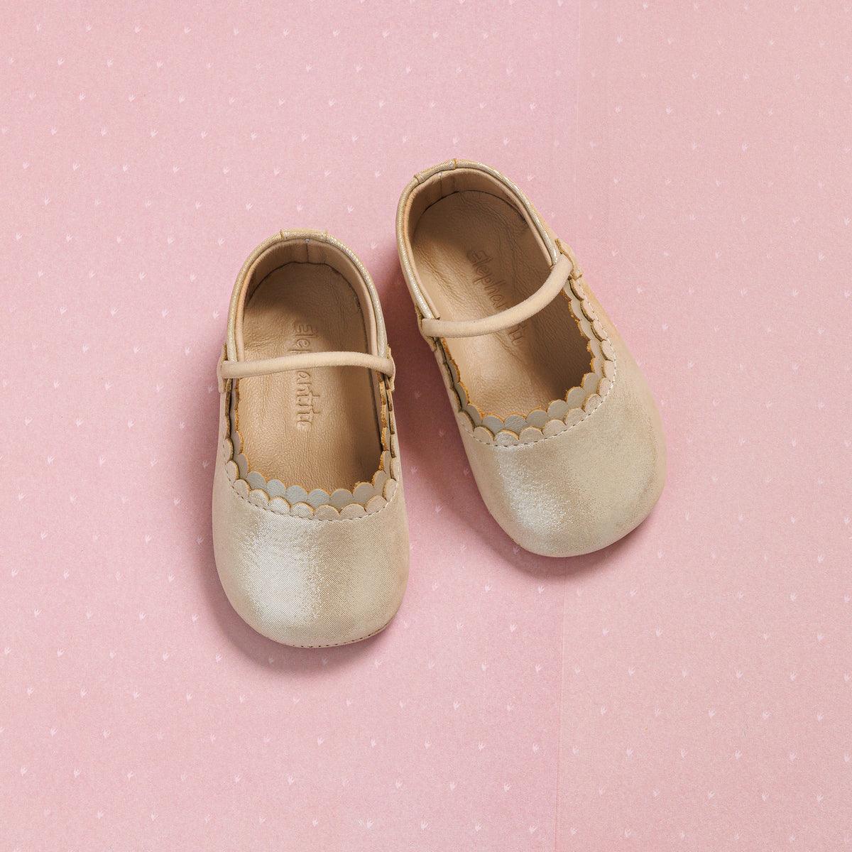 Cream-colored infant ballet shoes with scalloped edges and a Mary Jane strap on a pink polka dot background.