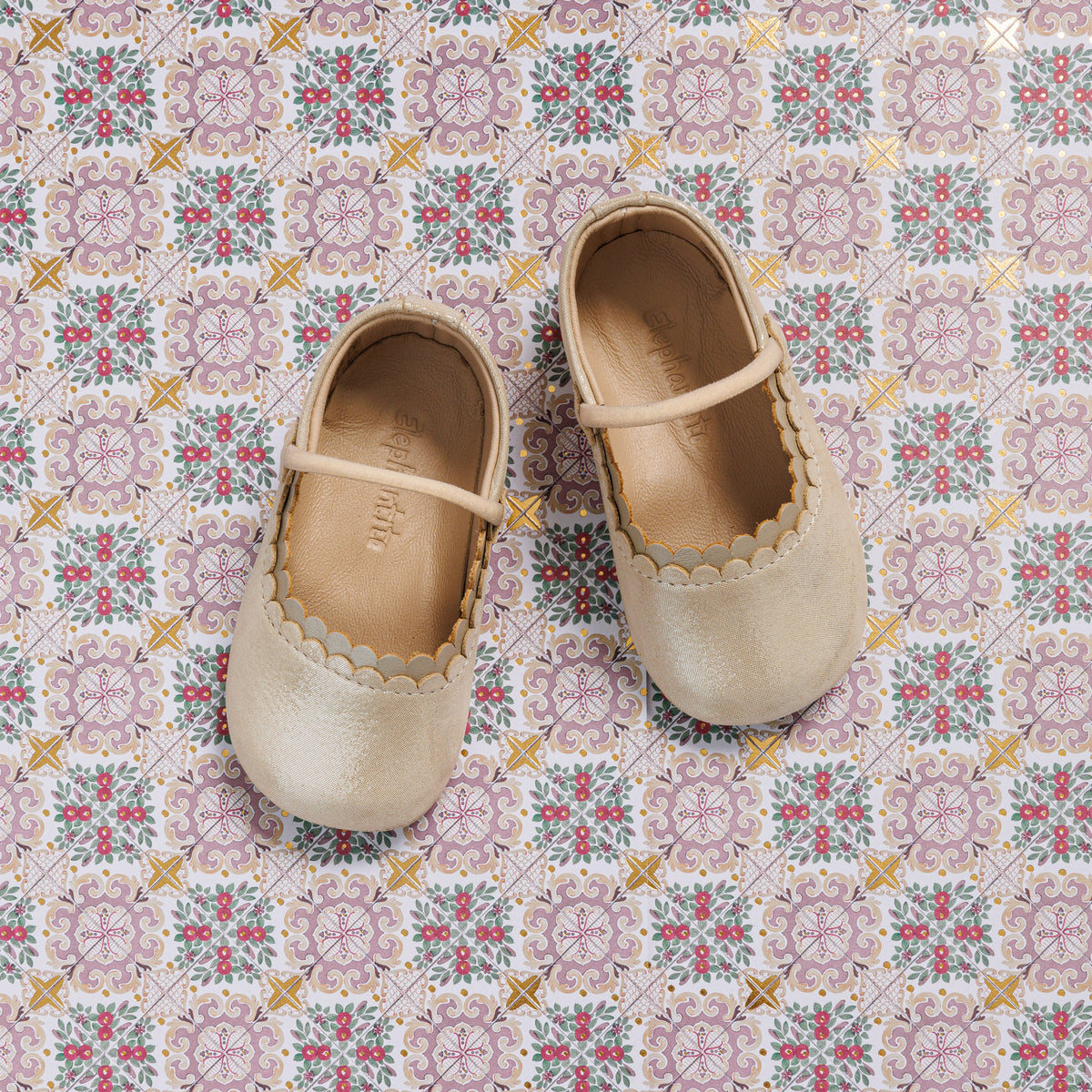 Cream-colored baby ballet flats with scalloped edges and a thin strap across the top, displayed on a patterned tile.