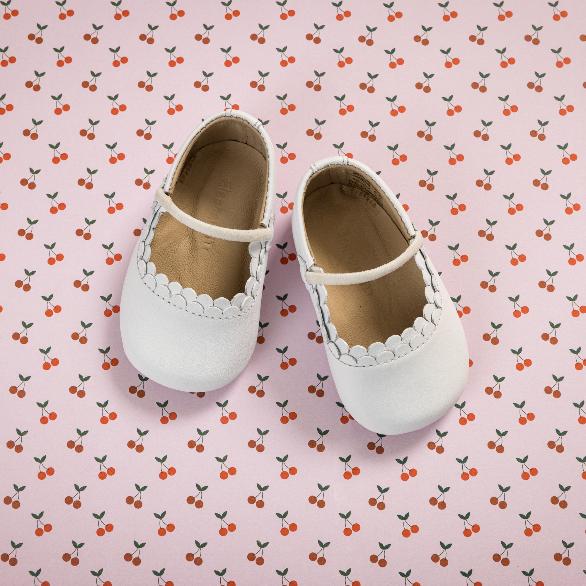 White baby ballet flats with scalloped edges and a single strap over the top on a pink cherry-patterned background.