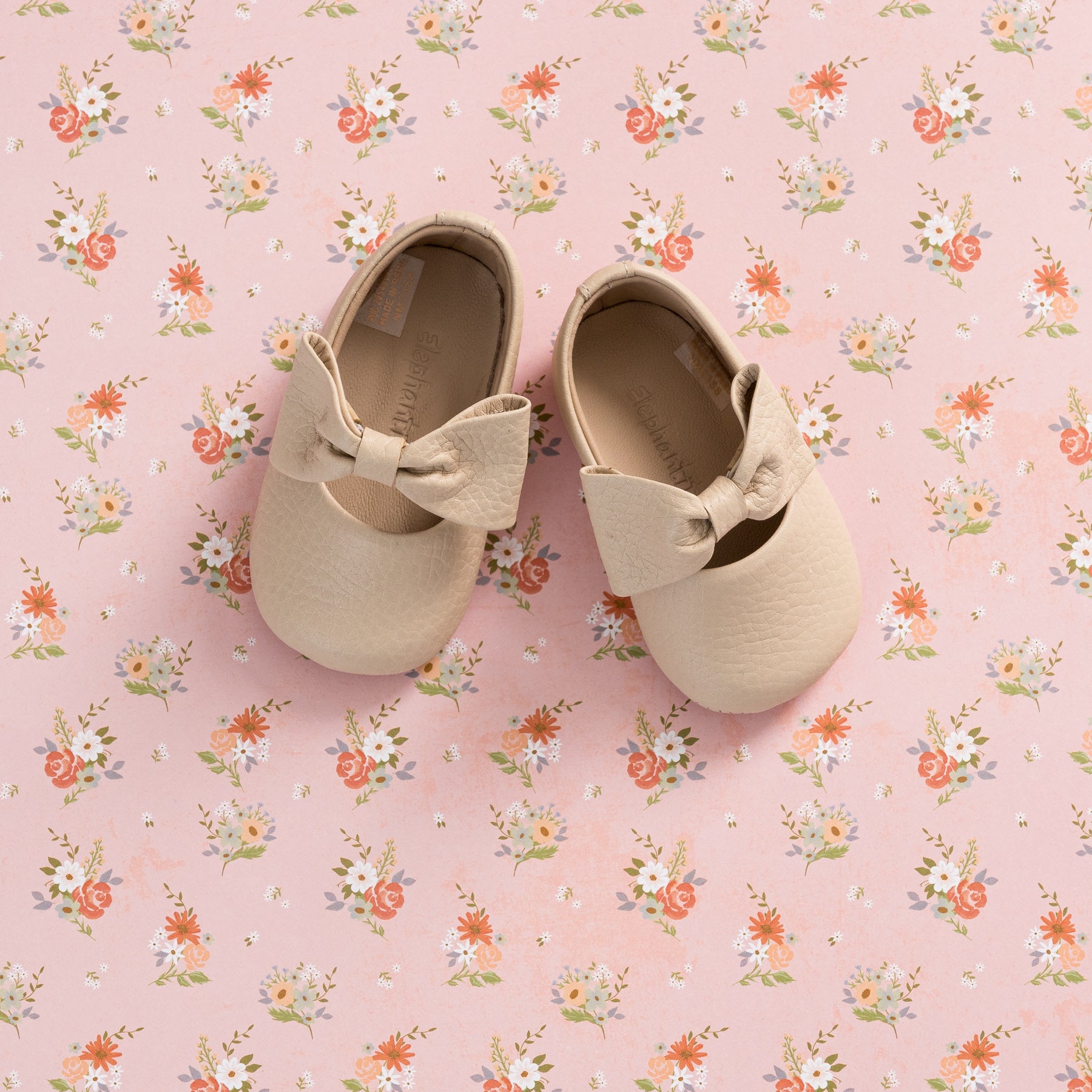 Cream-colored baby ballerina shoes with textured finish and large matching bow on pink floral background.