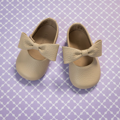 Cream-colored baby ballerina shoes with large textured bows and soft leather material on a patterned purple background.