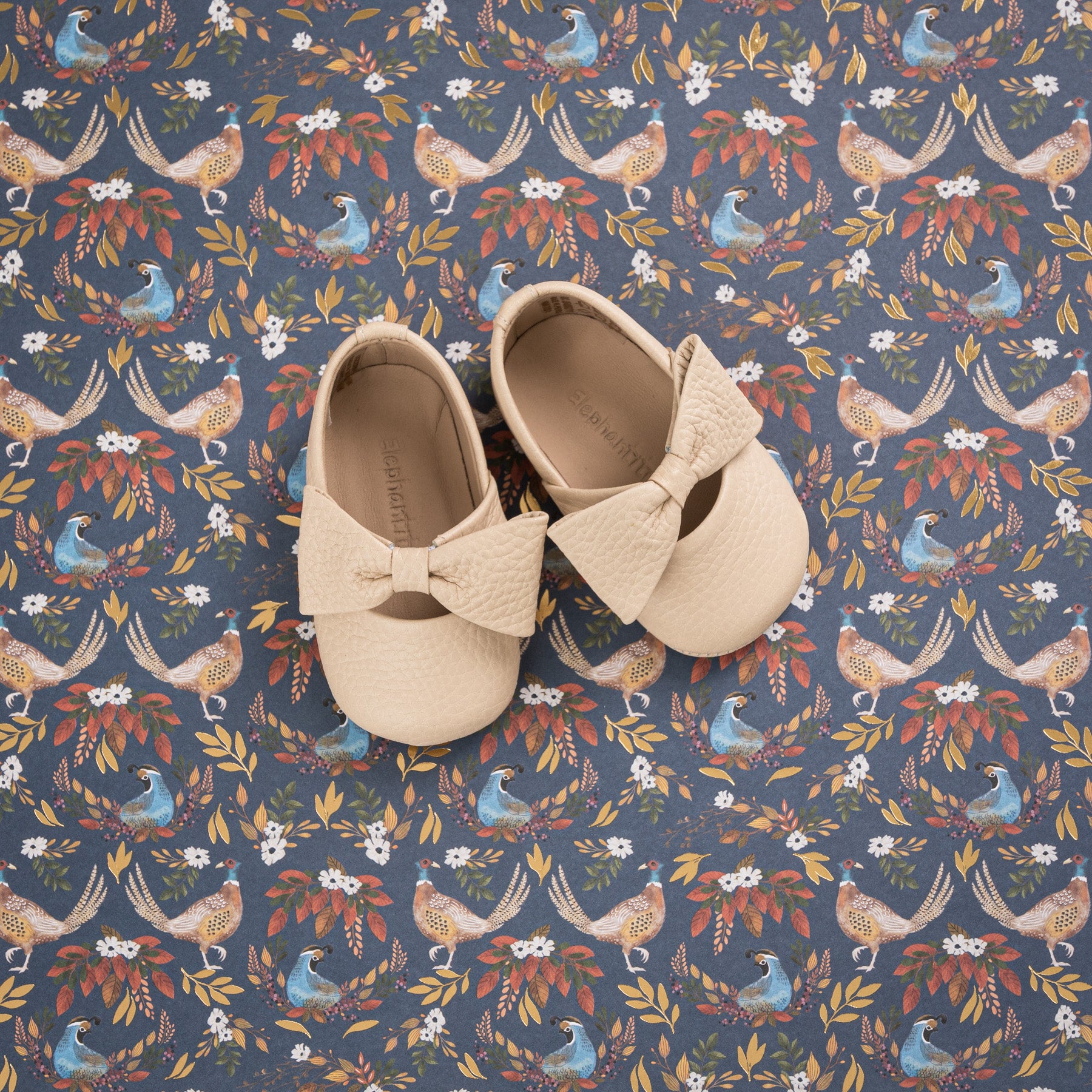 Cream-colored baby ballerina shoes with textured bow detail on floral patterned background.