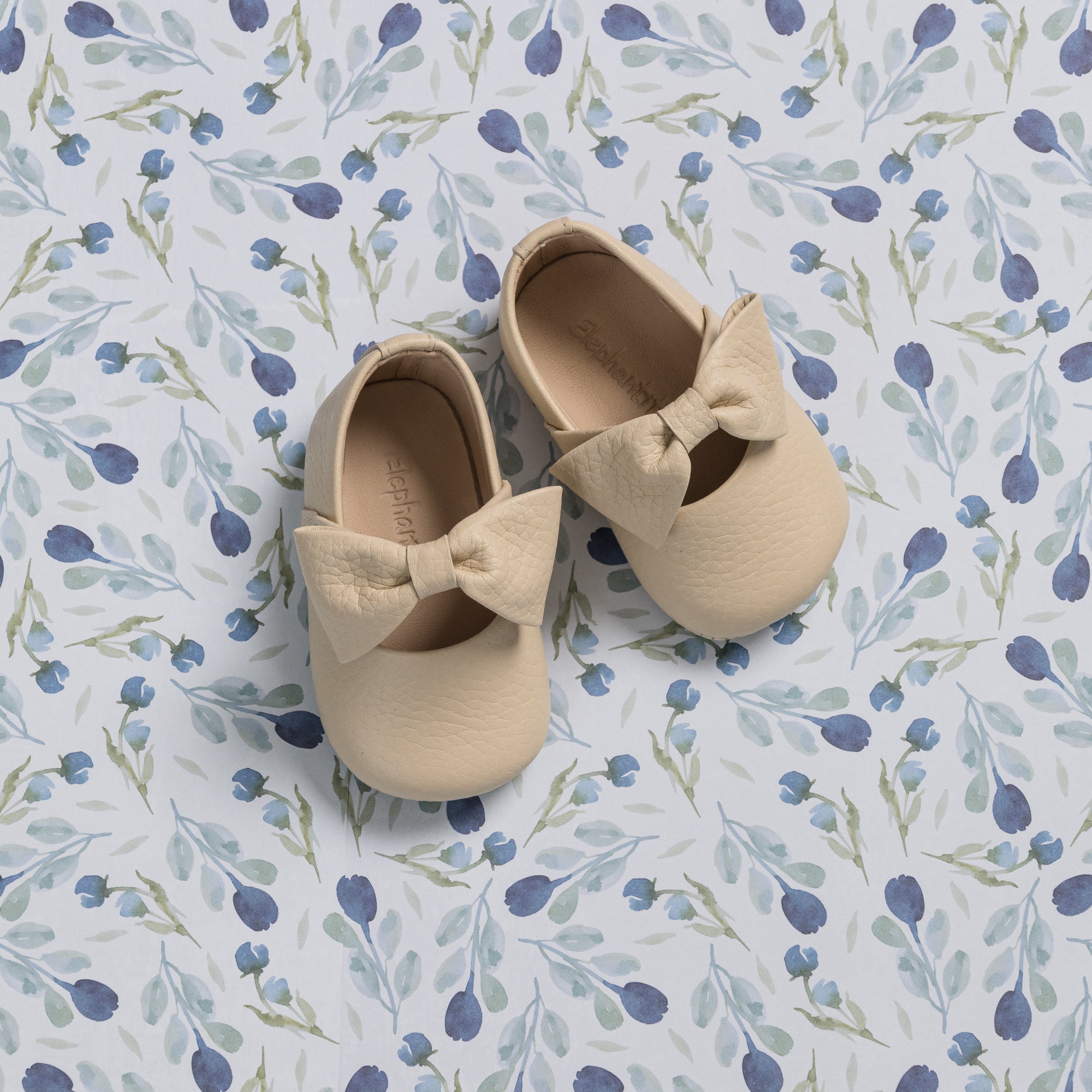 Cream-colored baby ballerina shoes with large fabric bows on soft floral background.