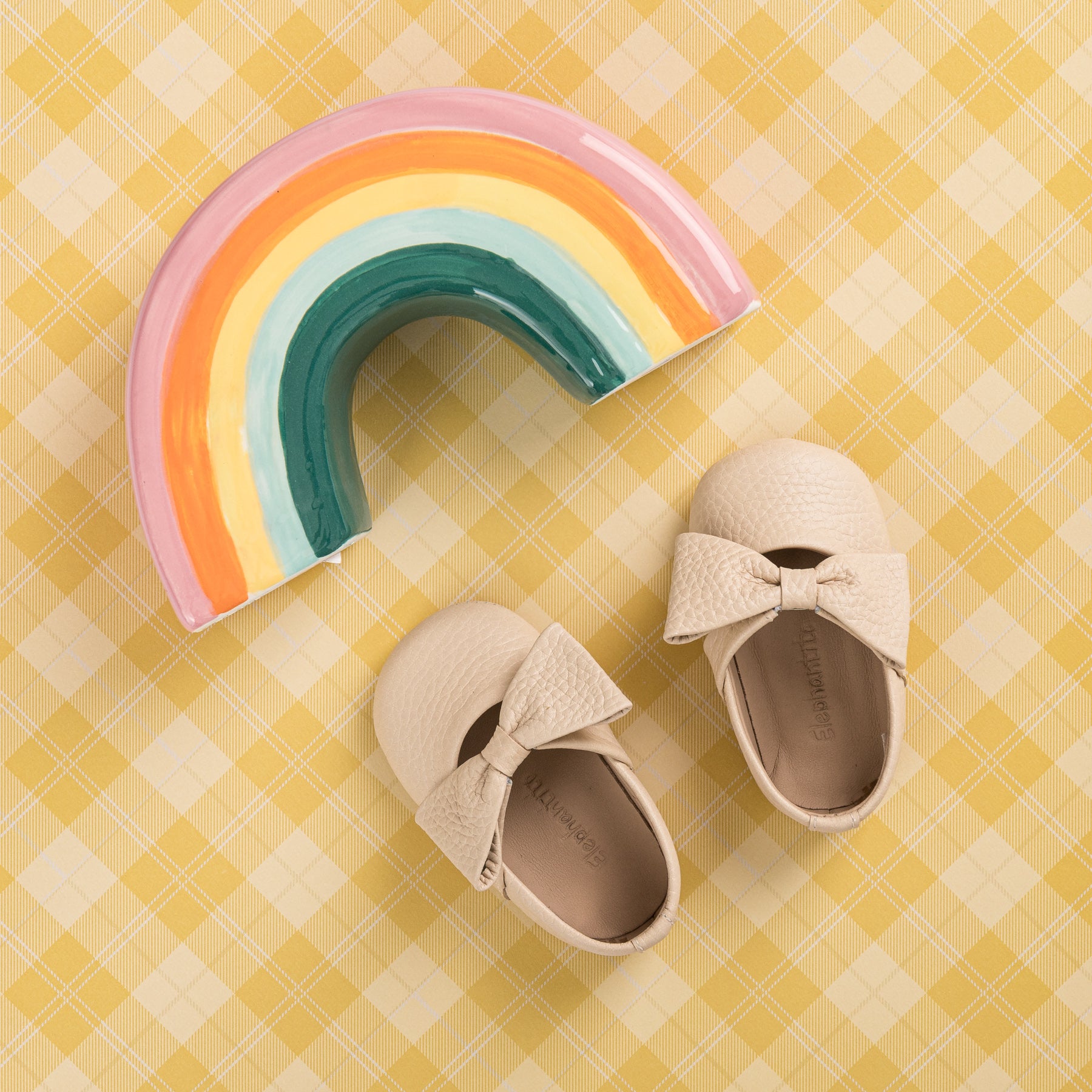 Cream-colored baby ballerina shoes with textured bows on a yellow checkered background, paired with a ceramic rainbow.