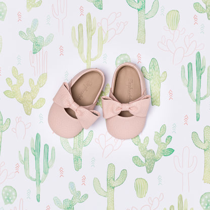 Soft pink infant ballerina shoes with decorative bow on a cactus-patterned background.