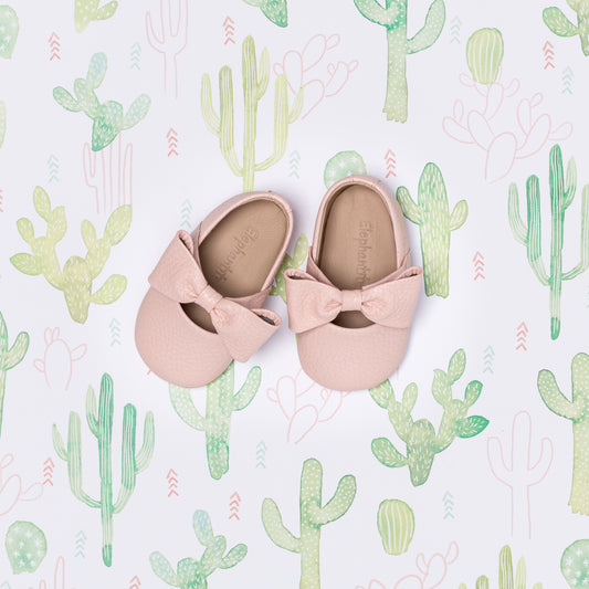 Soft pink infant ballerina shoes with decorative bow on a cactus-patterned background.