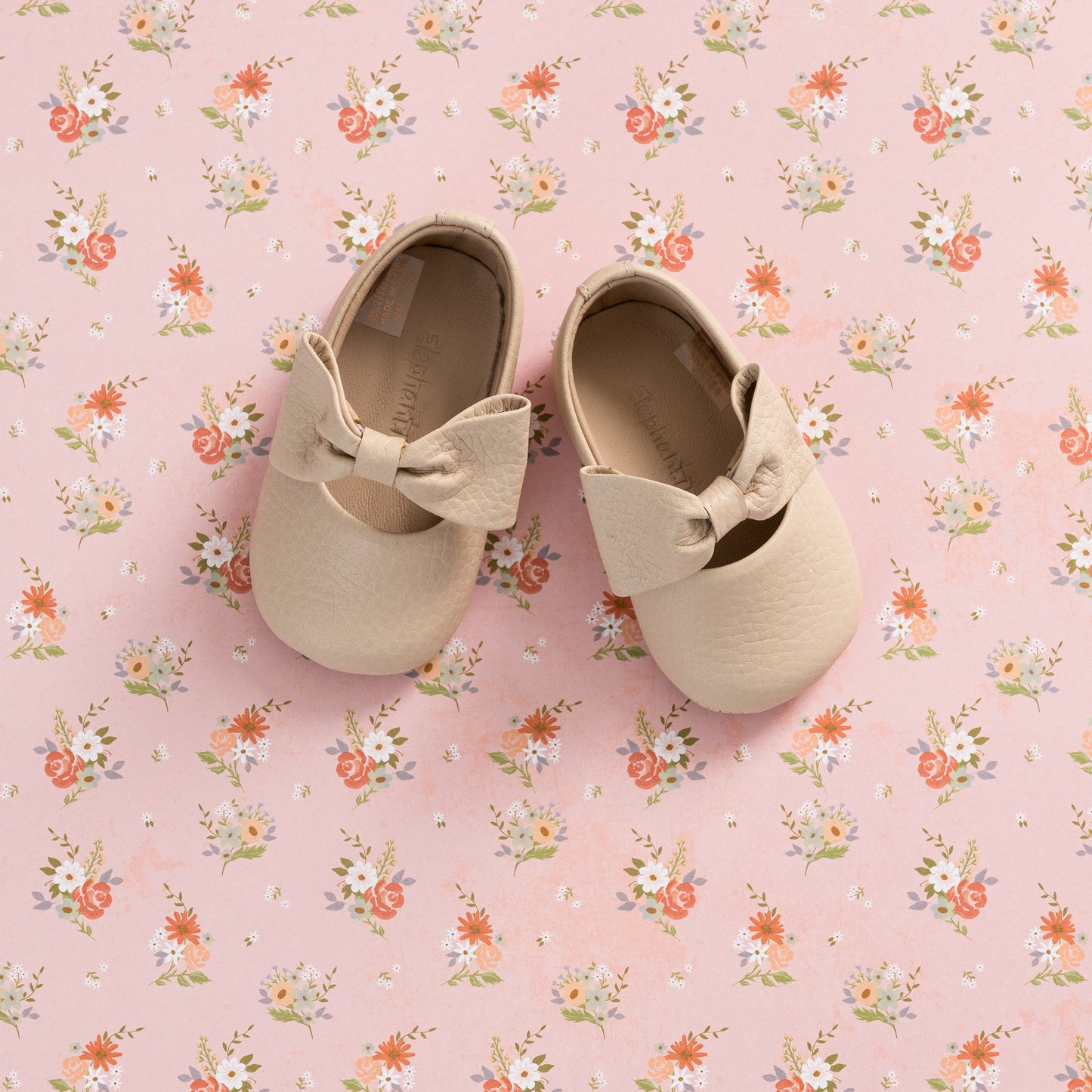 Soft pink baby shoes with textured finish and large matching bow on top, set on floral background.