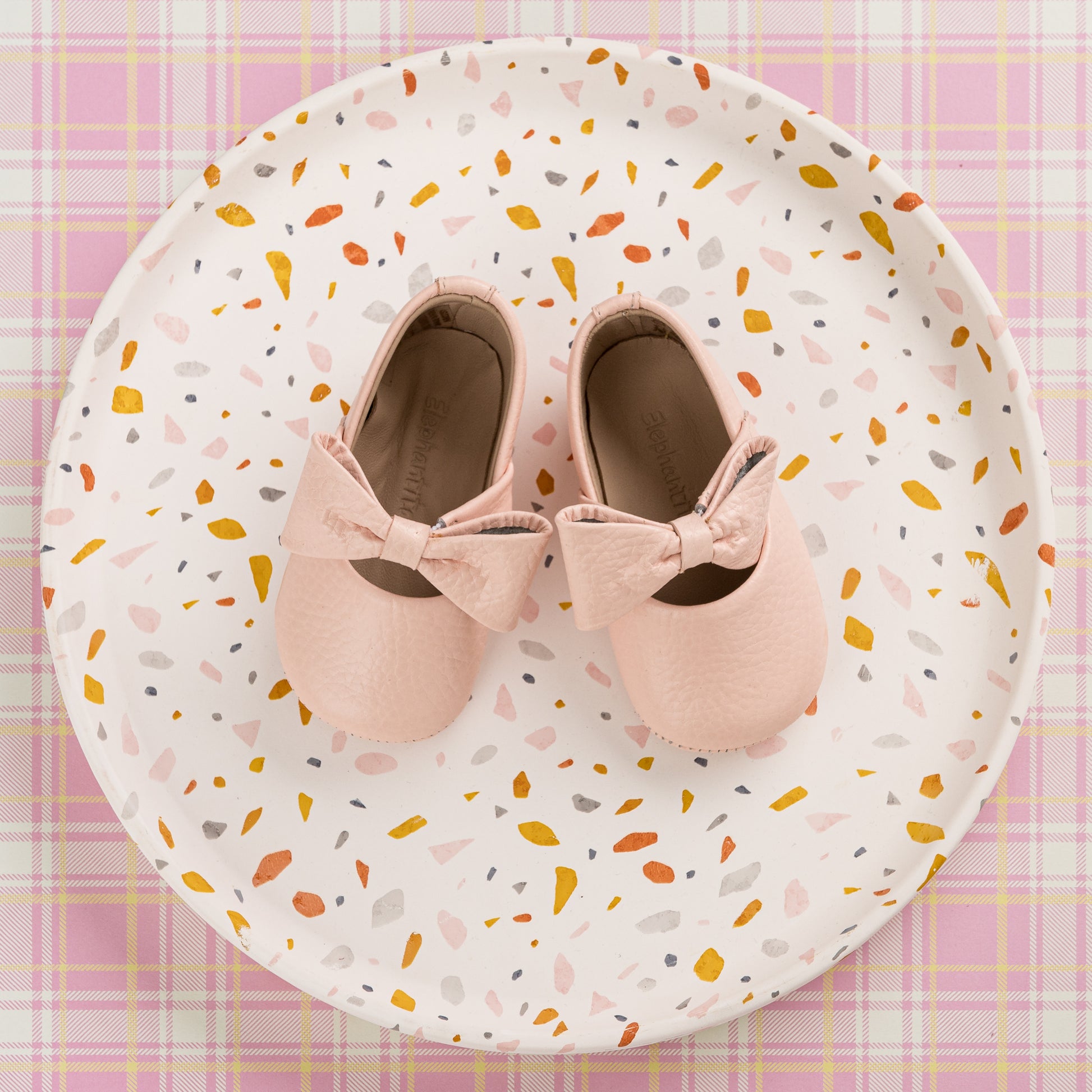 Soft pink baby ballerina shoes with decorative bows on top, displayed on a patterned plate.