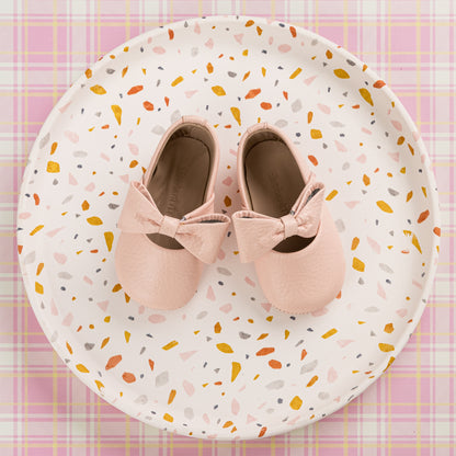 Soft pink baby ballerina shoes with decorative bows on top, displayed on a patterned plate.