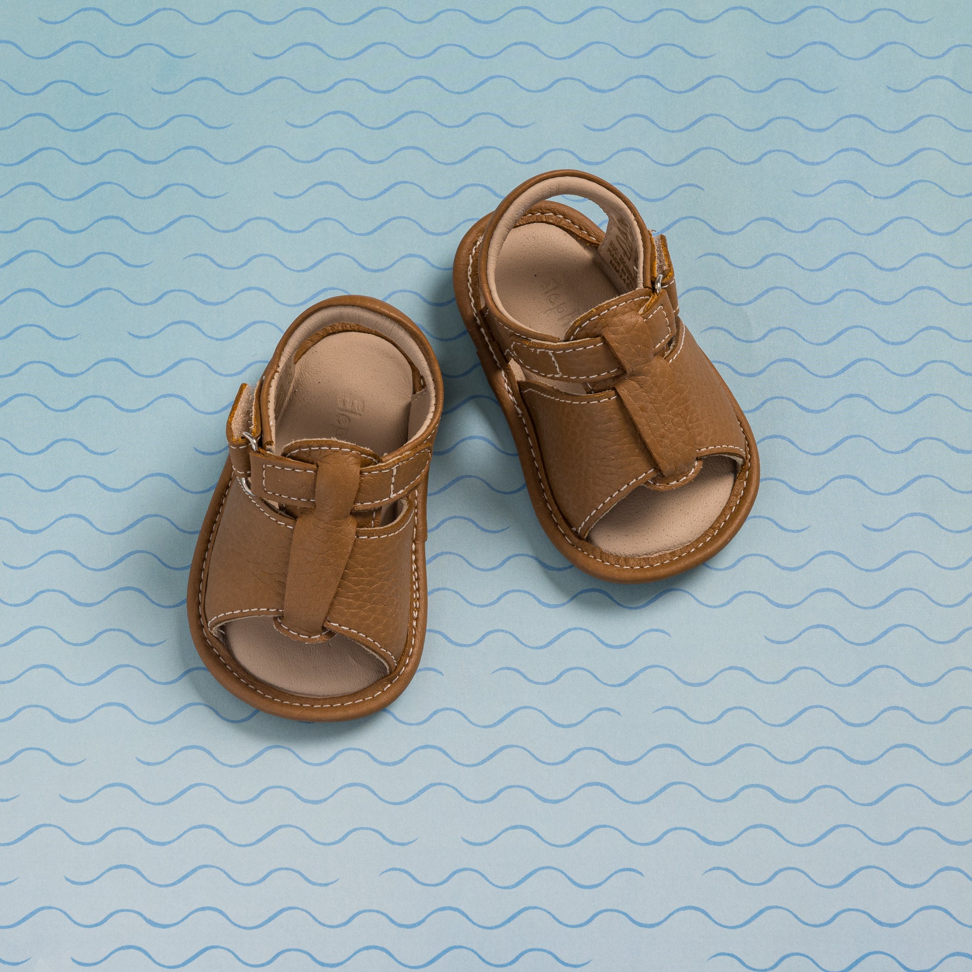 Caramel brown baby boy sandals with open toes and Velcro straps on blue wavy background.