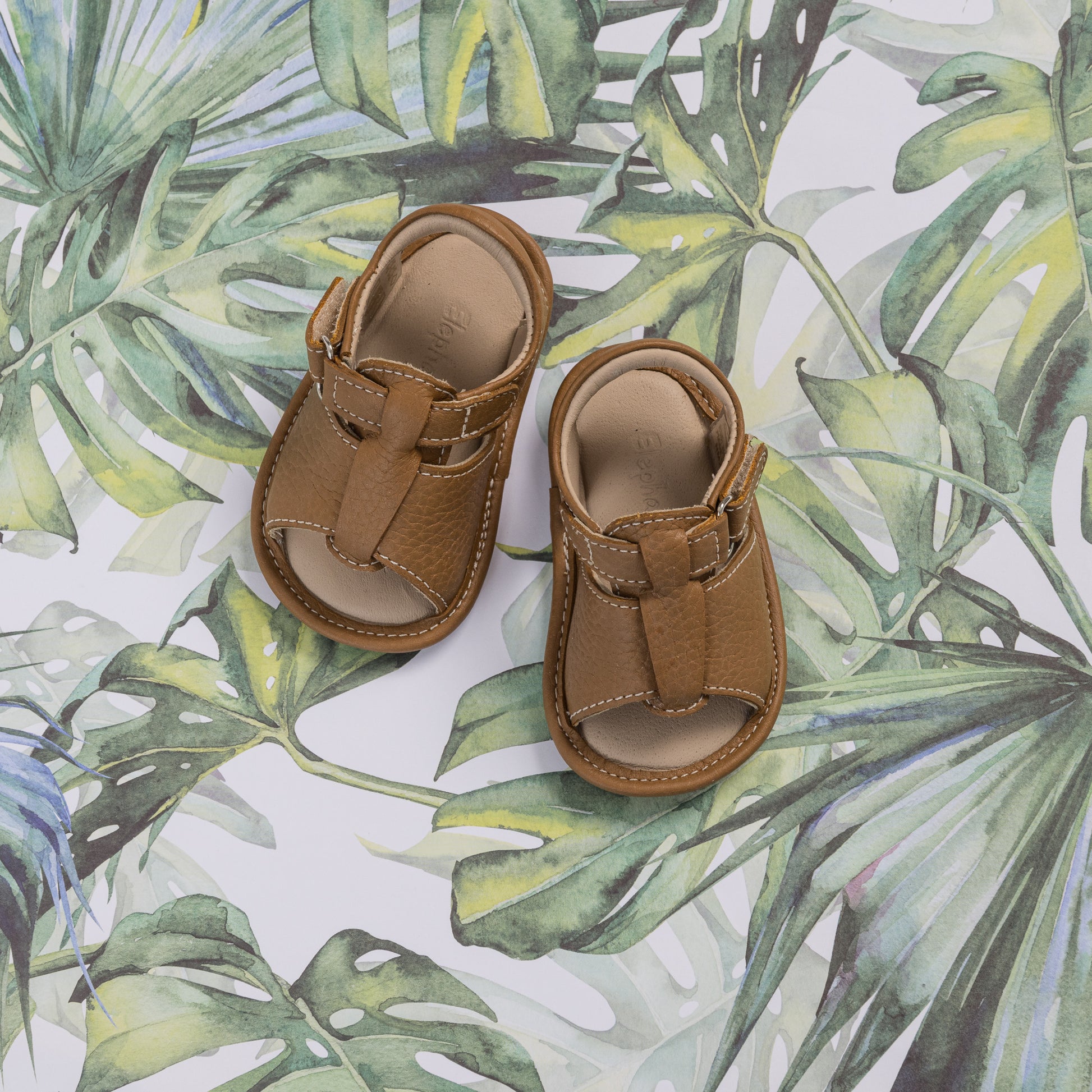 Caramel brown baby boy sandals with adjustable straps and soft soles on leafy background.