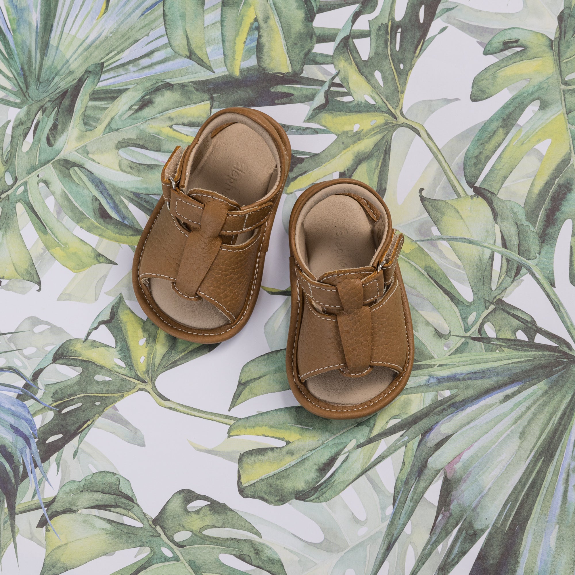 Caramel brown baby boy sandals with adjustable straps and soft soles on leafy background.