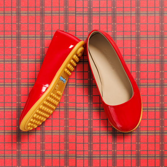 Glossy red toddler ballerina flats with soft beige insoles and textured tan rubber soles on red plaid background.
