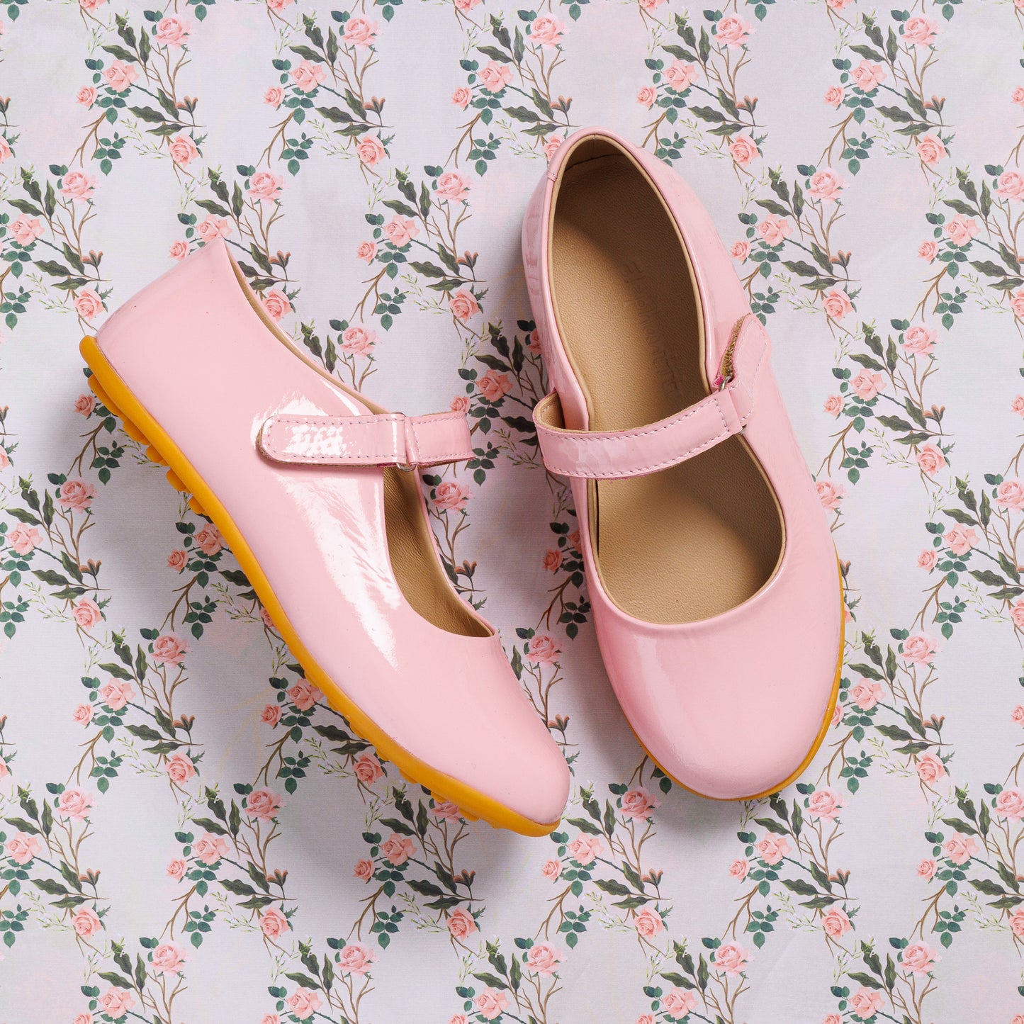 Glossy light pink toddler Mary Jane shoes with a strap and rubber sole on floral background.