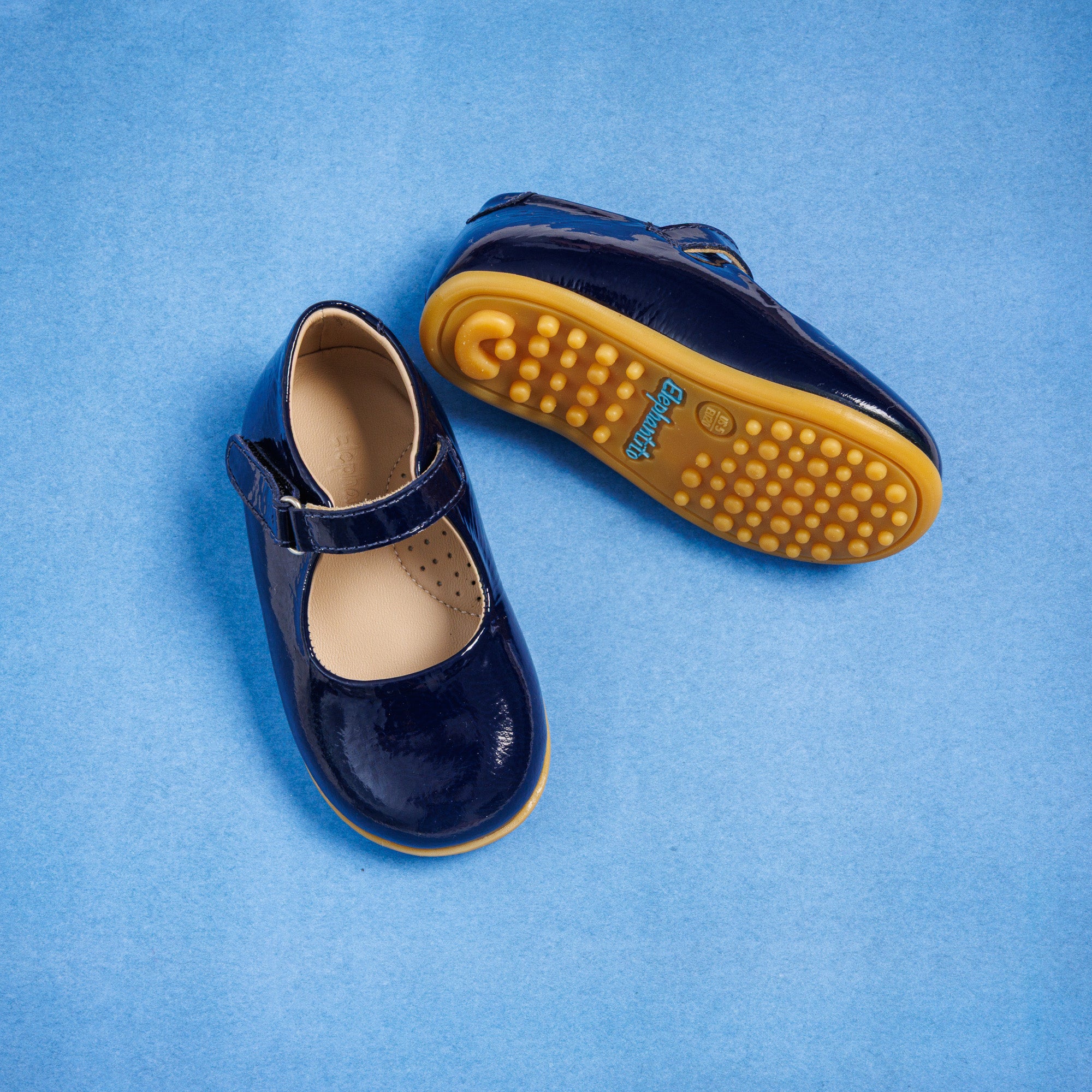 Glossy navy blue toddler Mary Jane shoes with velcro strap and textured tan soles on blue background.