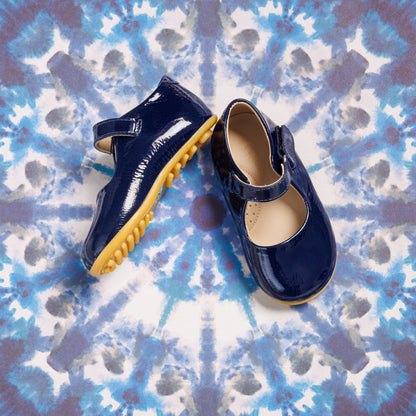 Glossy navy blue toddler Mary Jane shoes with tan rubber soles on tie-dye background.