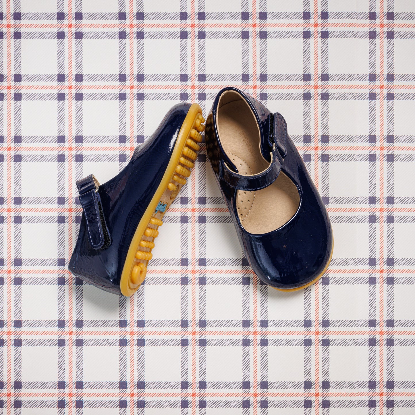 Navy blue patent leather toddler Mary Jane shoes with rounded toes and yellow rubber soles on plaid background.