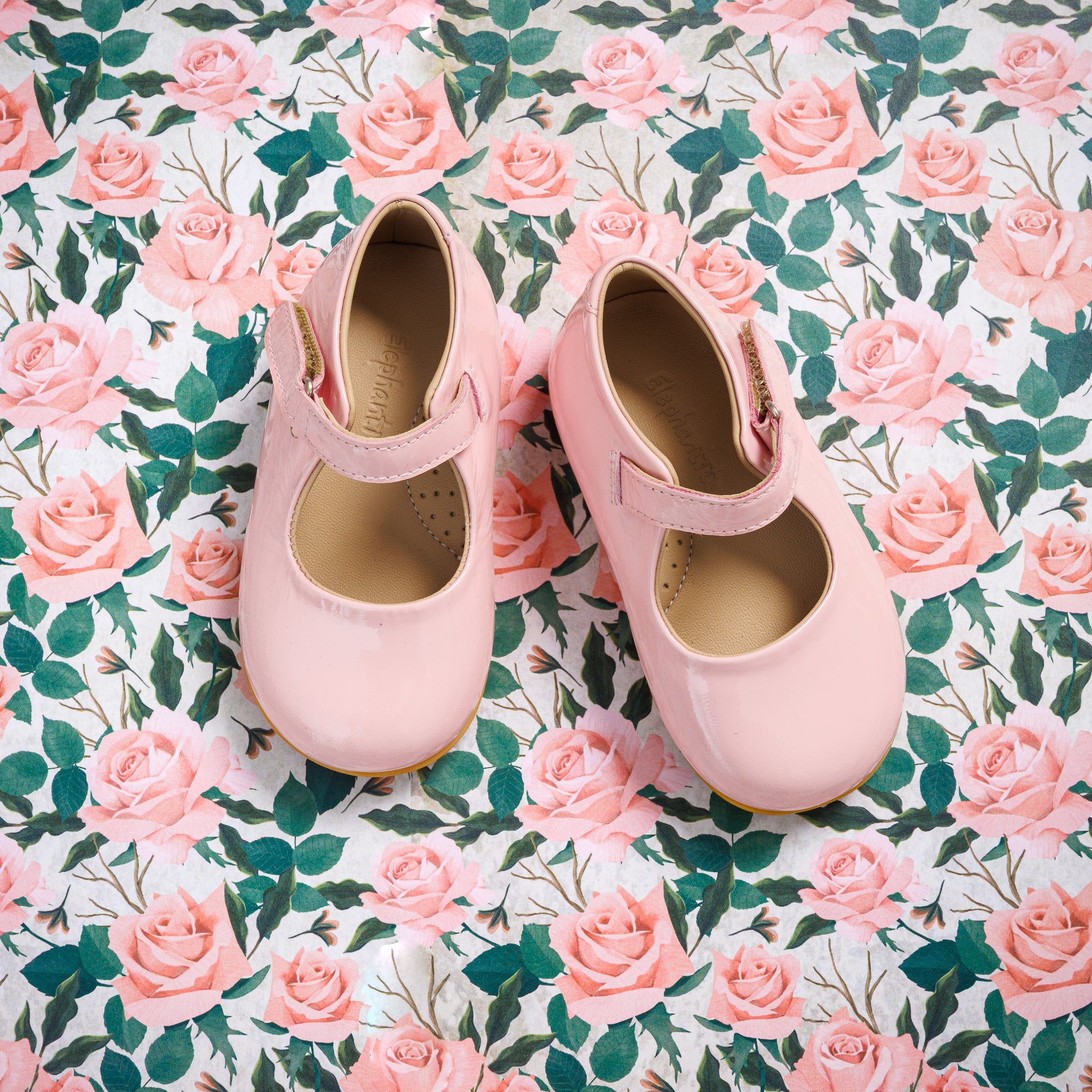 Pink toddler Mary Jane shoes with single straps on floral rose-patterned fabric background.