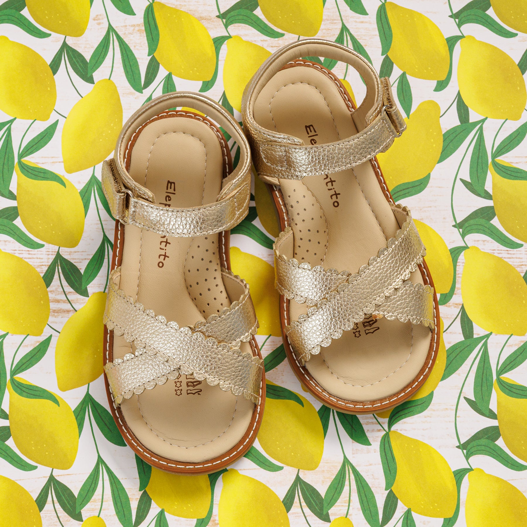 Elephantito - Gold metallic toddler sandals with crisscross straps and adjustable ankle closures on lemon-print background.