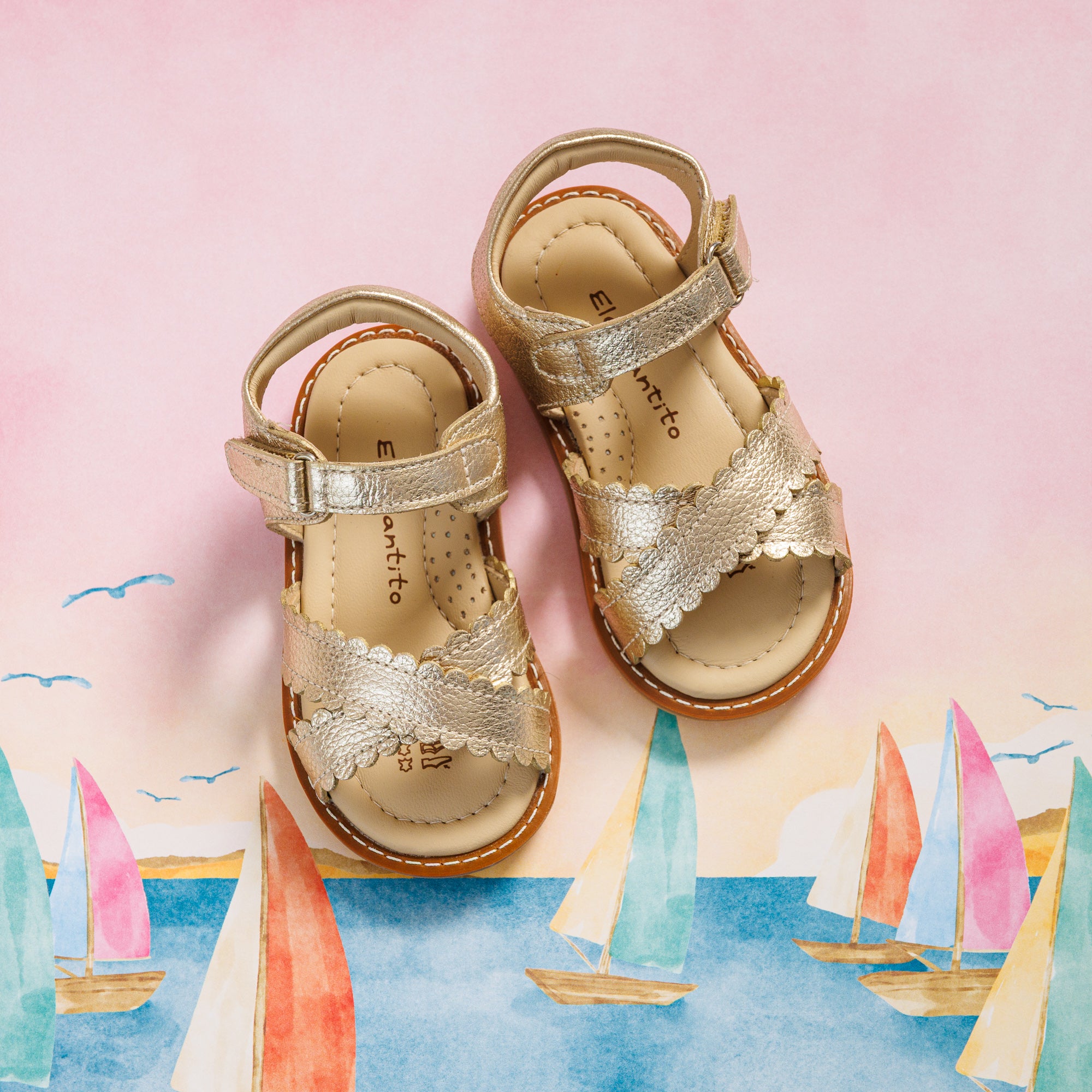 Elephantito - Toddler gold metallic sandals with scalloped straps and adjustable ankle buckle on painted sailboat background.