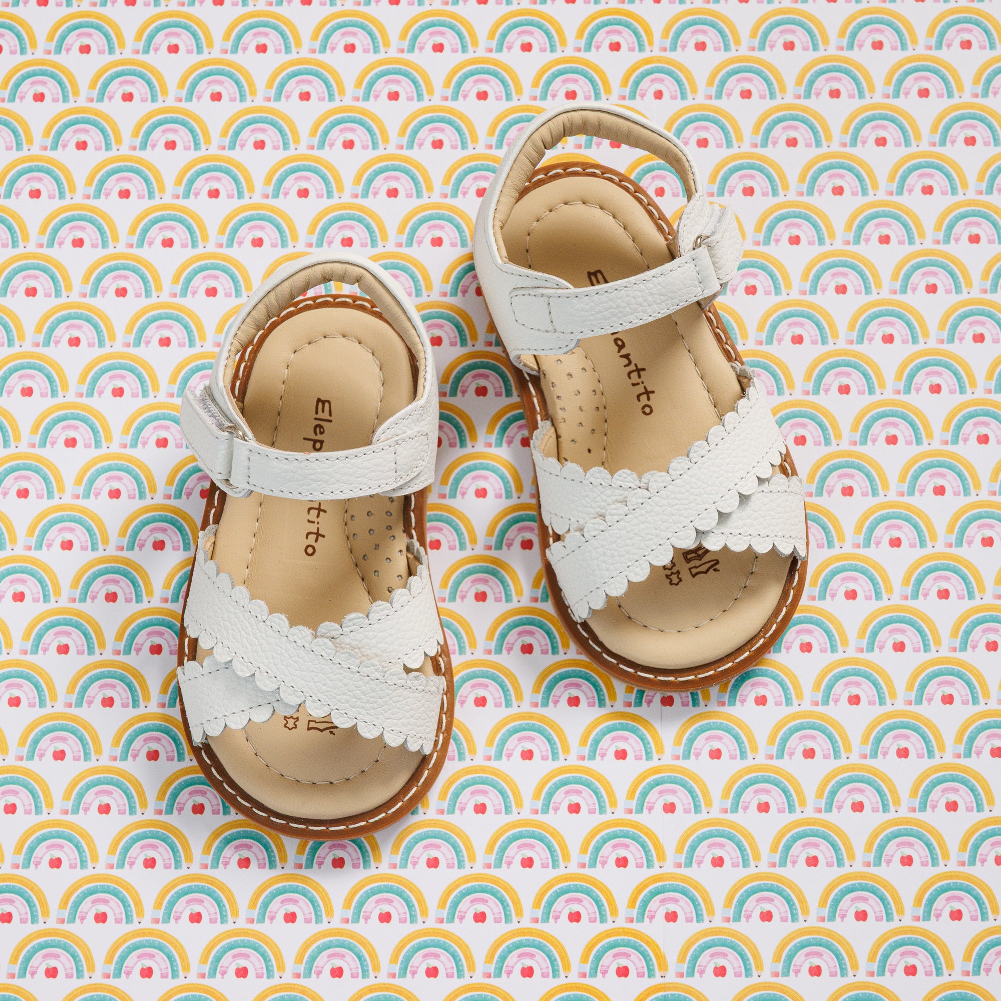 Elephantito - White toddler sandals with scalloped straps, open toes, and cushioned tan insoles on rainbow background.