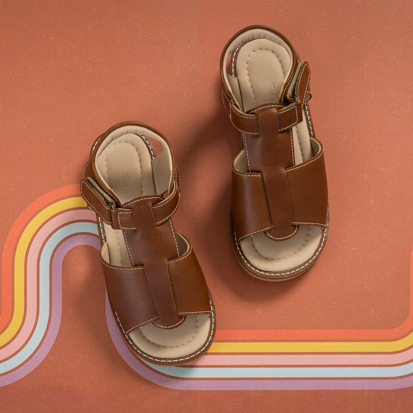 Natural brown leather boy sandals with cushioned insoles and adjustable straps on rust background with rainbow stripe design.