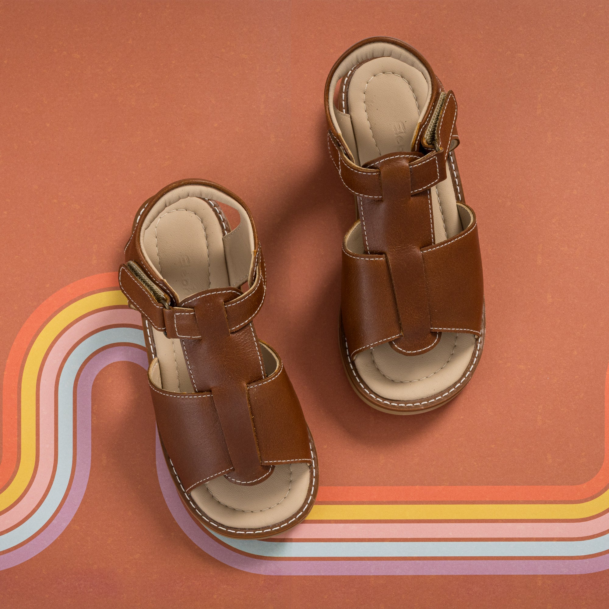 Natural brown leather boy sandals with cushioned insoles and adjustable straps on rust background with rainbow stripe design.