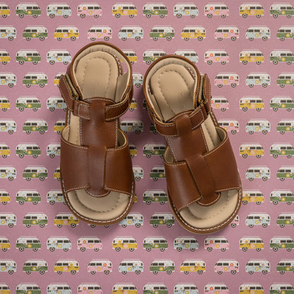 Brown leather toddler sandals with adjustable straps and beige cushioned soles on pink background with colorful vans pattern.