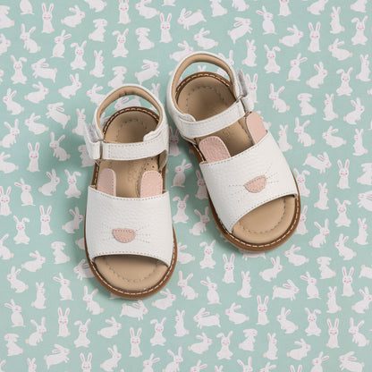 White toddler sandals with bunny cutout detail and adjustable ankle straps on bunny-patterned background.