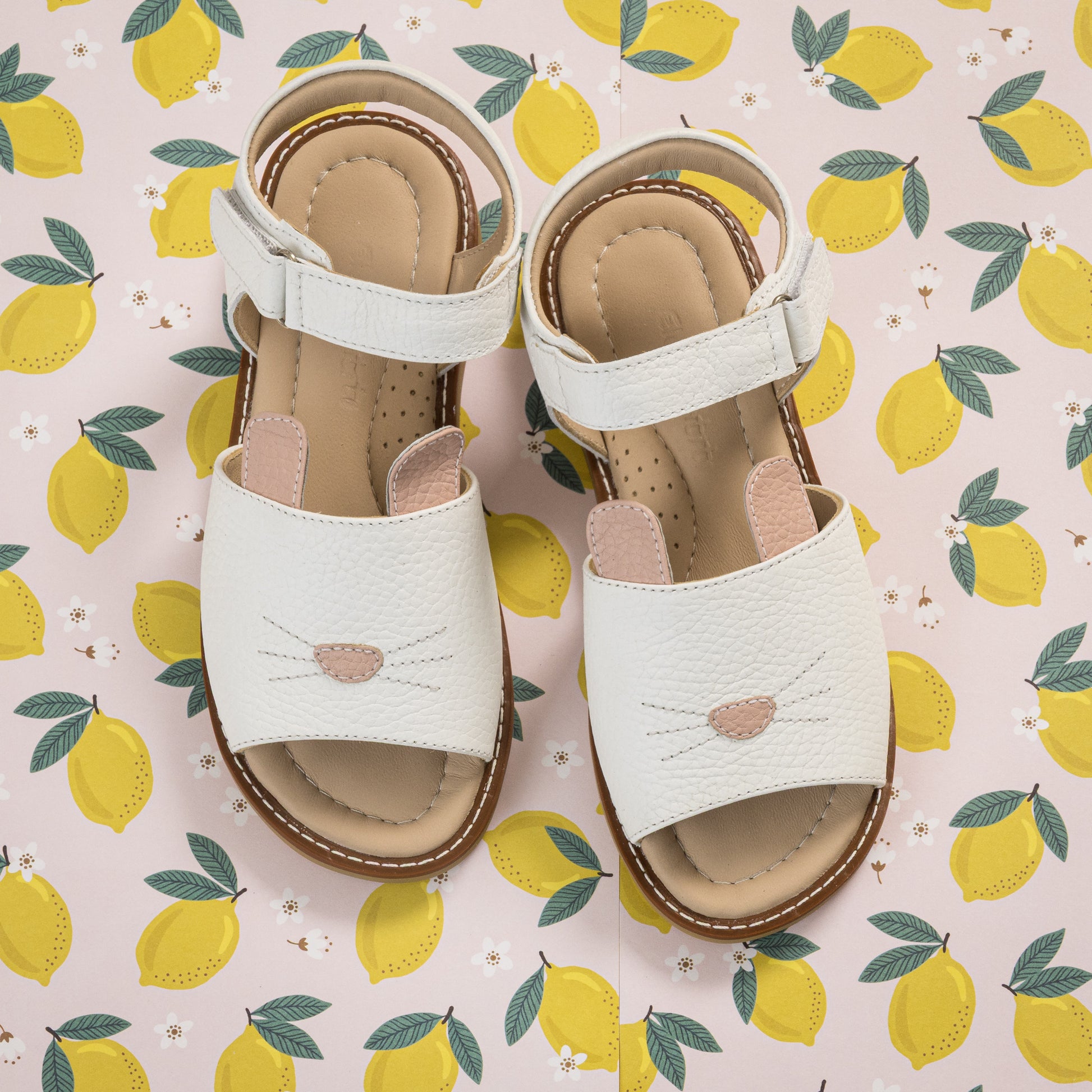 White toddler sandals with bunny face design and adjustable ankle straps on lemon-patterned background.