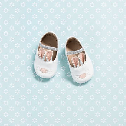 White baby slippers with soft bunny ears and pink embroidered nose on light blue floral background.