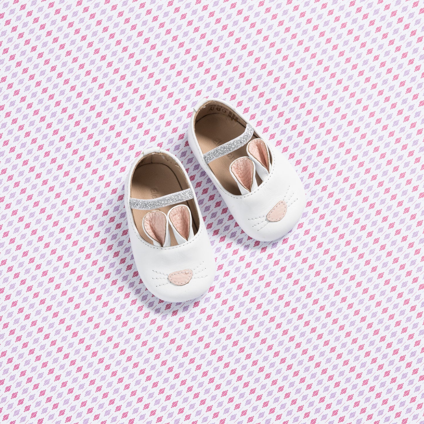 White baby slippers shaped like bunnies with pink accents and soft soles on a patterned background.