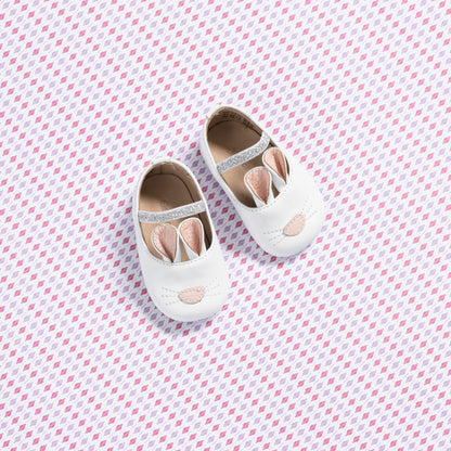 White baby slippers shaped like bunnies with pink accents and soft soles on a patterned background.