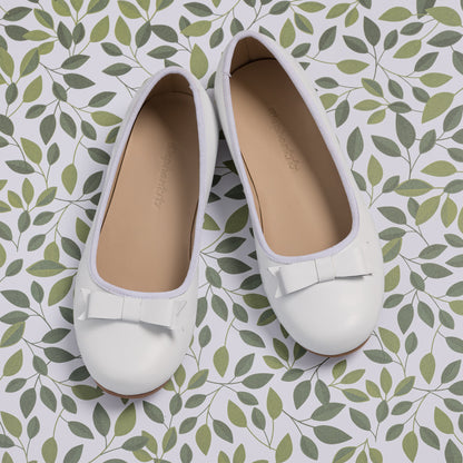 White toddler ballet flats with rounded toes and small decorative bows on top, set on leafy patterned surface.