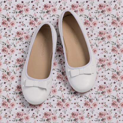 White toddler ballet flats with rounded toes and decorative bows on top, set on floral fabric background.