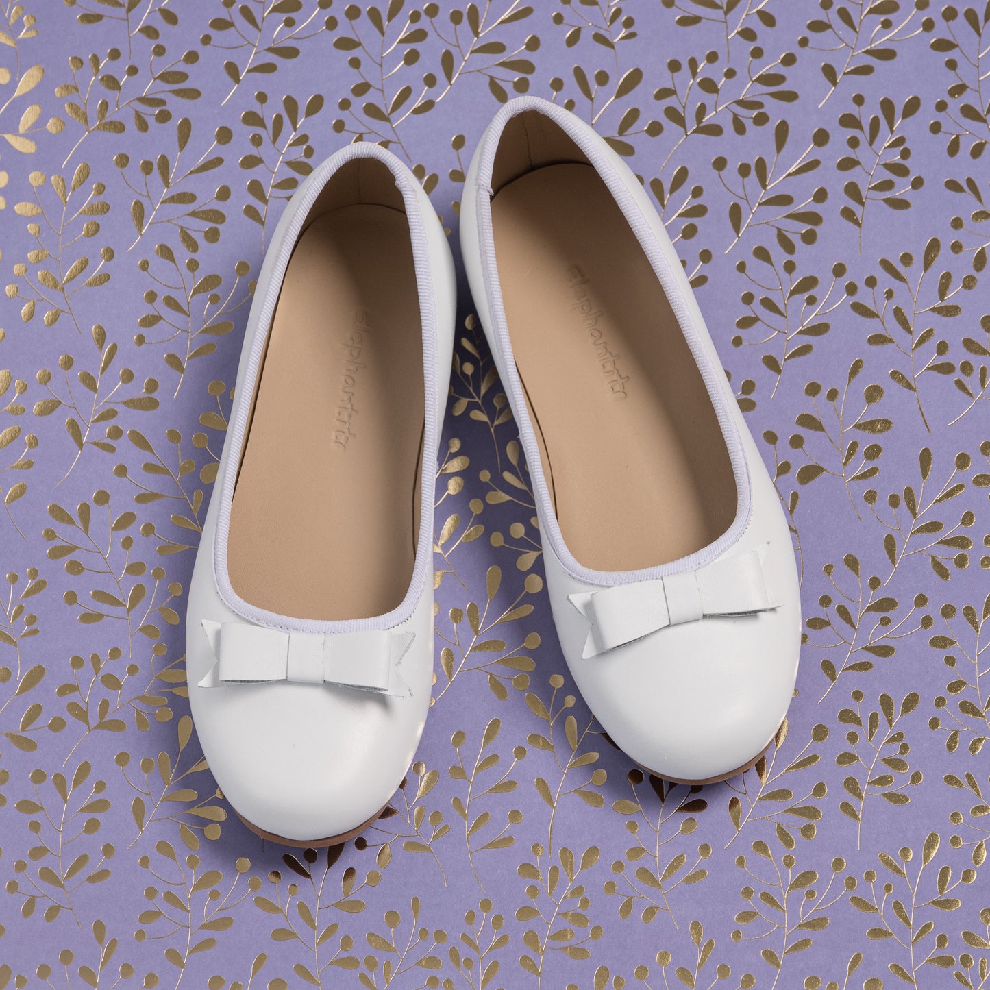 White children's ballerina flats with rounded toes and small decorative bows on top, tan insoles.