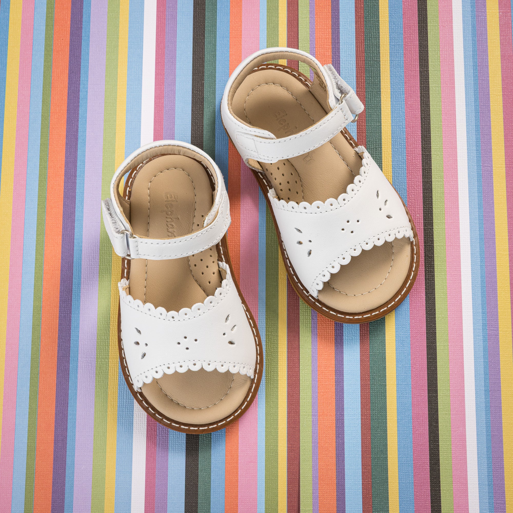 White toddler sandals with scalloped edges and ankle straps on colorful striped background.