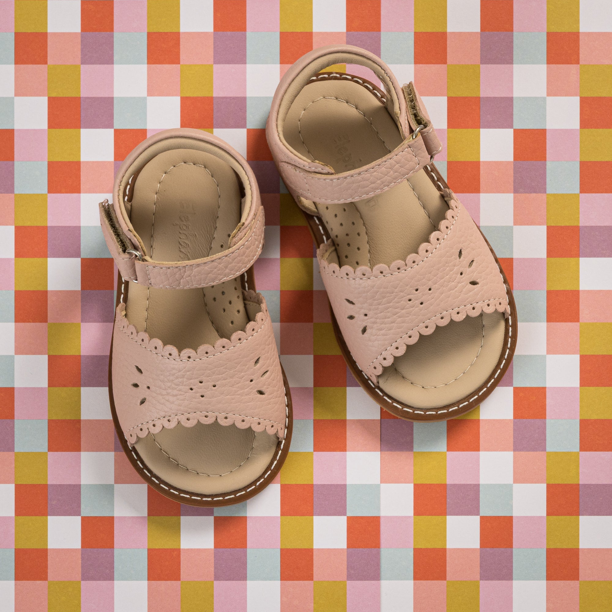 Pink toddler sandals with scalloped edges and ankle straps on a colorful checkered background.