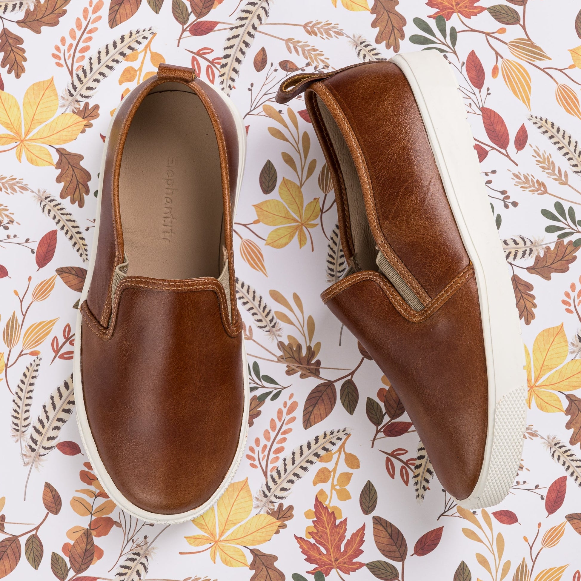 Natural brown leather slip-on kids shoes with white rubber soles and elastic side panels on autumn leaf print background.