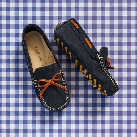 Elephantito - Navy blue suede boys' driver loafers with orange bow and rubber pebble sole detailing on gingham background.