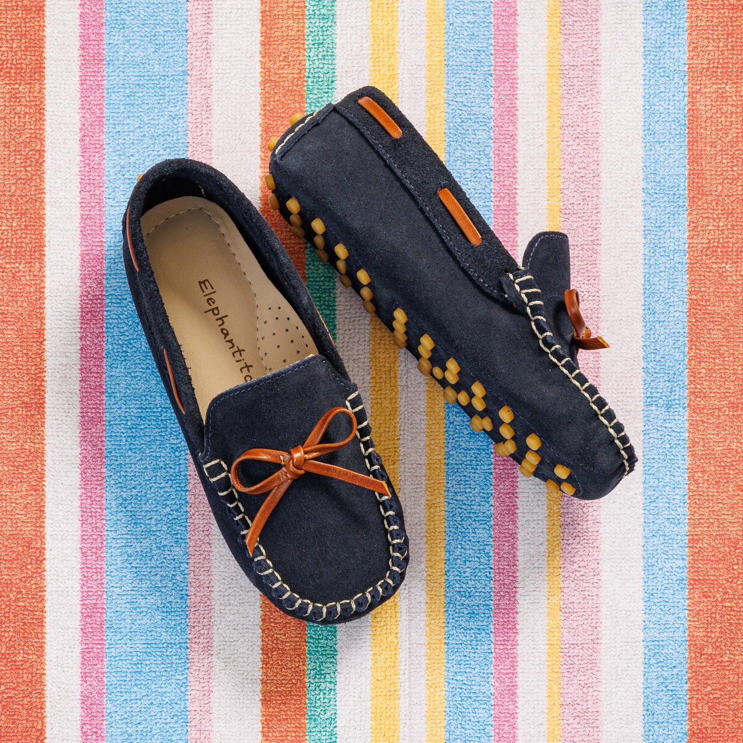 Elephantito - Navy blue suede boys' driver loafers with orange lace and yellow rubber sole grips on striped background.
