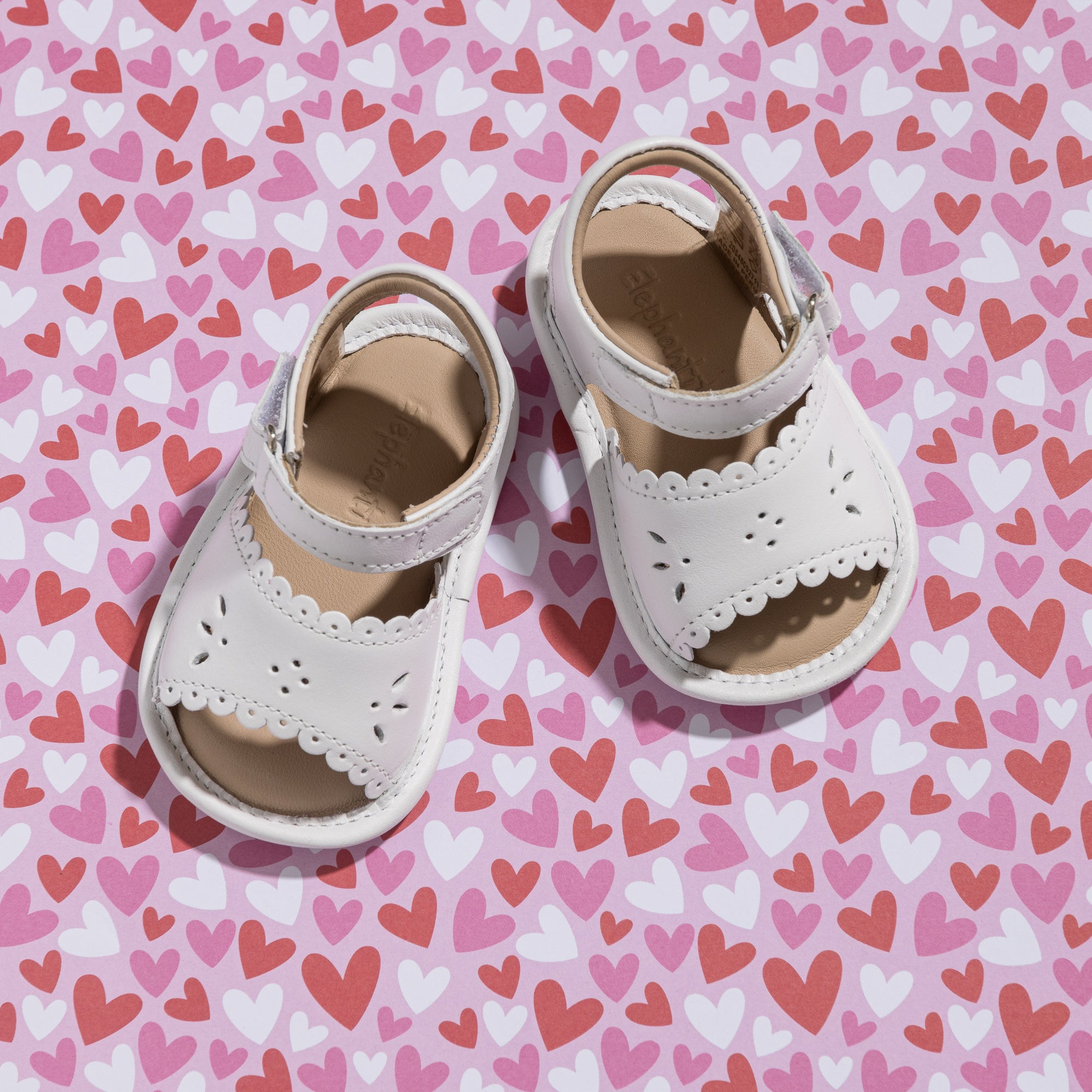White toddler sandals with scalloped leather straps and velcro ankle closures on a pink heart-patterned surface.