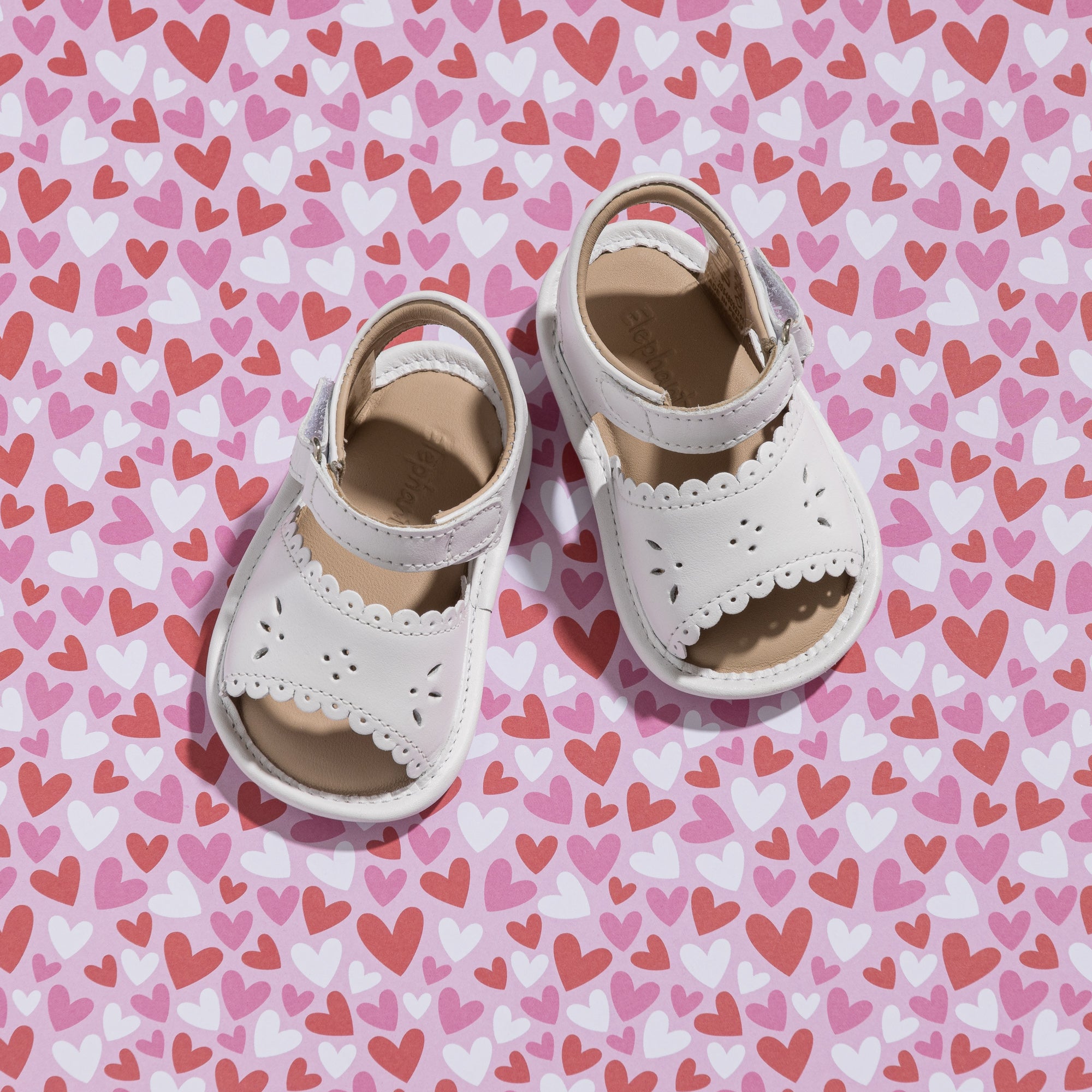 White toddler sandals with scalloped edges and ankle straps on heart-patterned background.