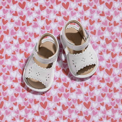 White toddler sandals with scalloped edges and ankle straps on heart-patterned background.