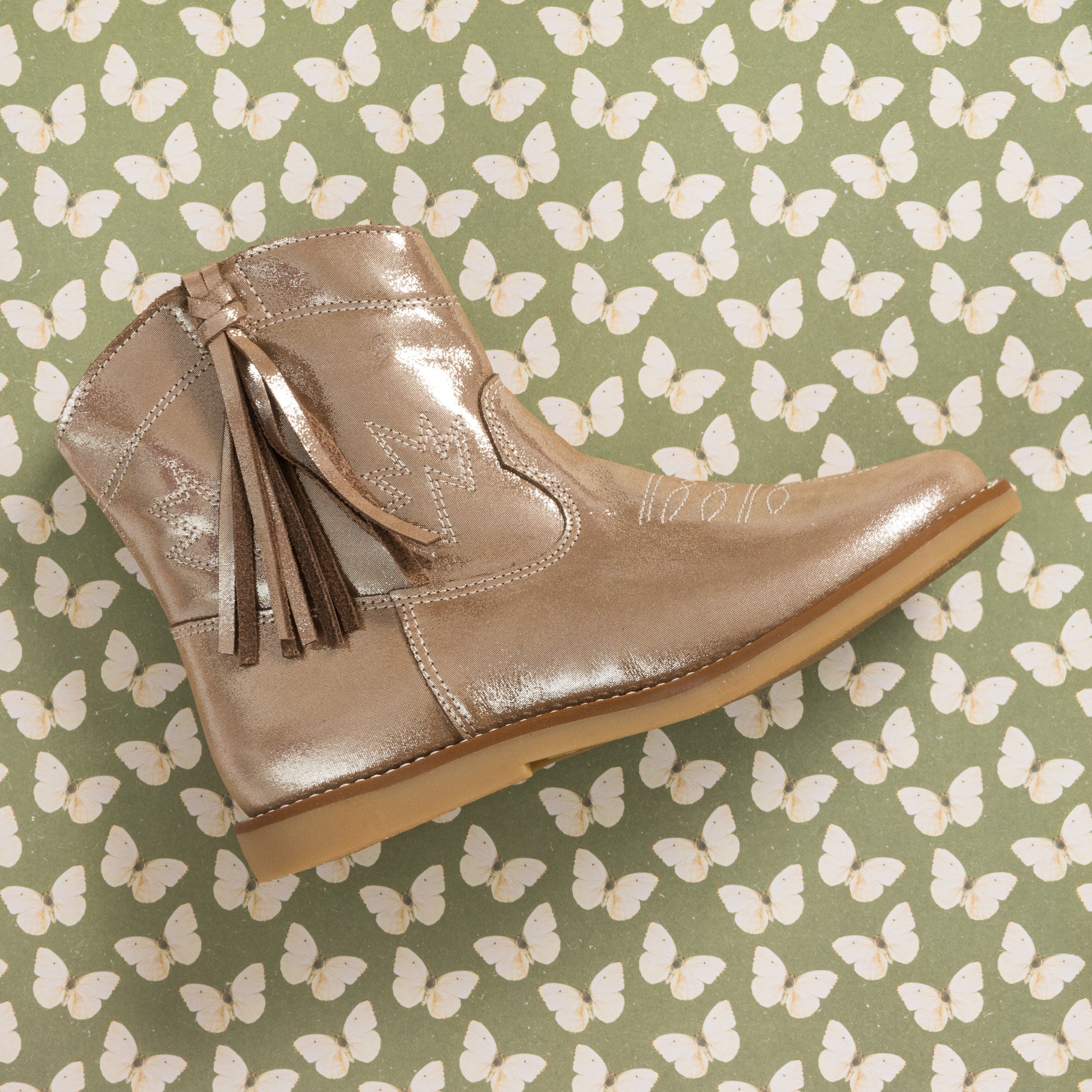 Metallic blush suede toddler boot with side tassels and stitched detailing on butterfly-patterned background.