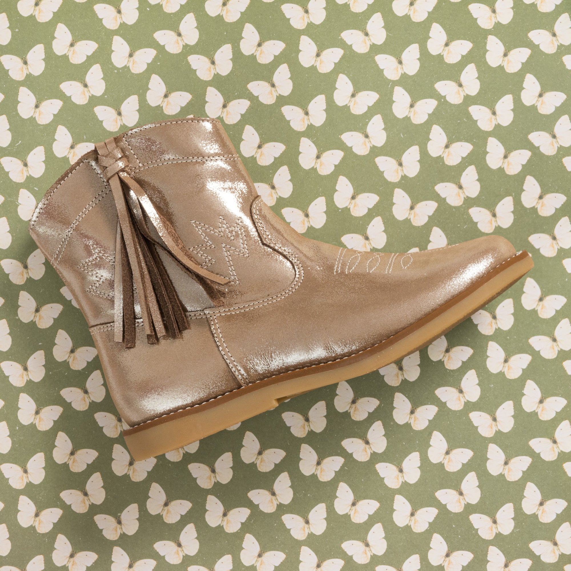 Metallic blush suede toddler boot with side tassels and stitched detailing on butterfly-patterned background.