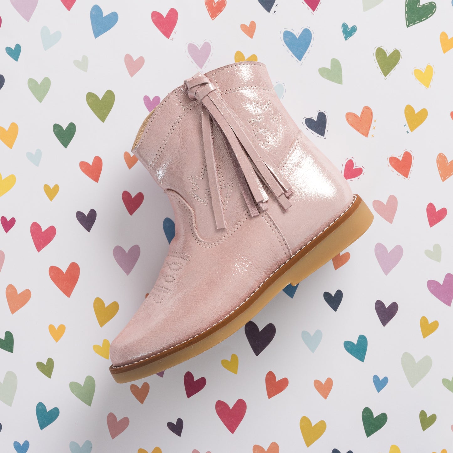 Metallic pink suede toddler boot with tassel zipper and light brown sole against colorful heart background.