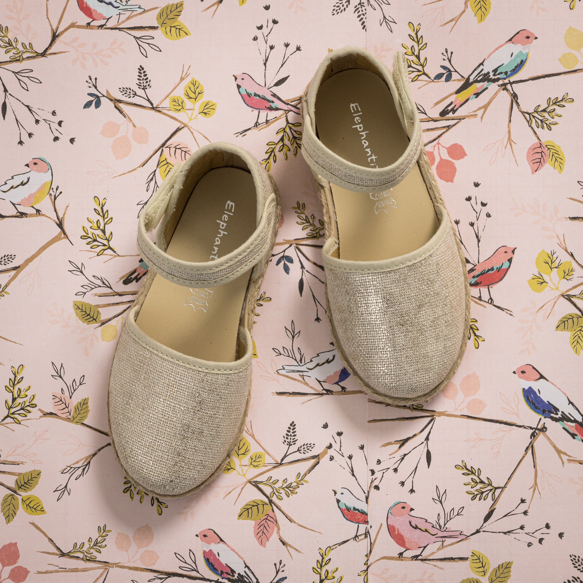 Metallic white linen espadrille with ankle strap and braided jute sole on floral background.